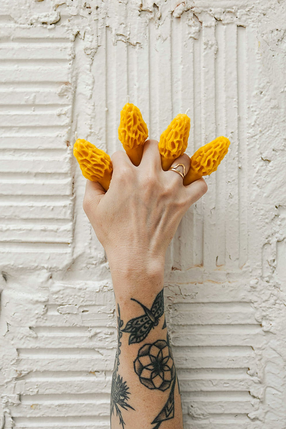 A hand holding a beeswax candle shaped like a morel mushroom, with a textured white wall in the background.