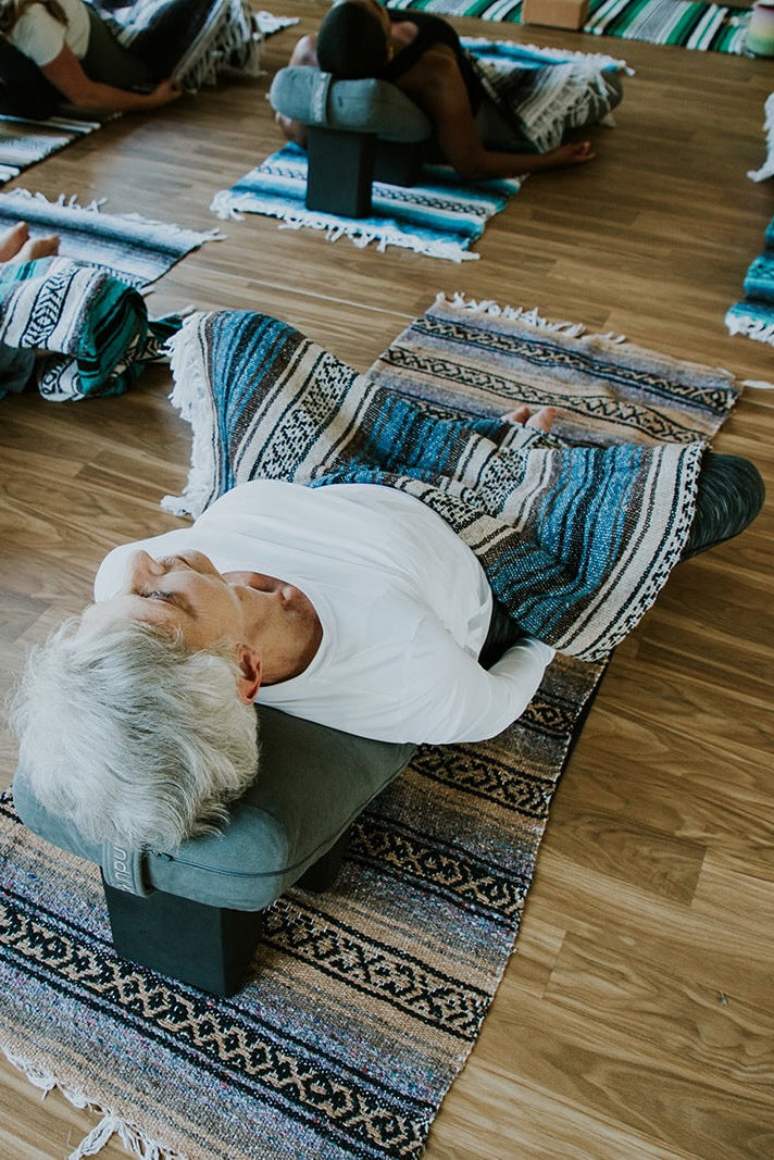 Woman in a restorative yoga pose with other people in a class with blankets, bolsters, and blocks.