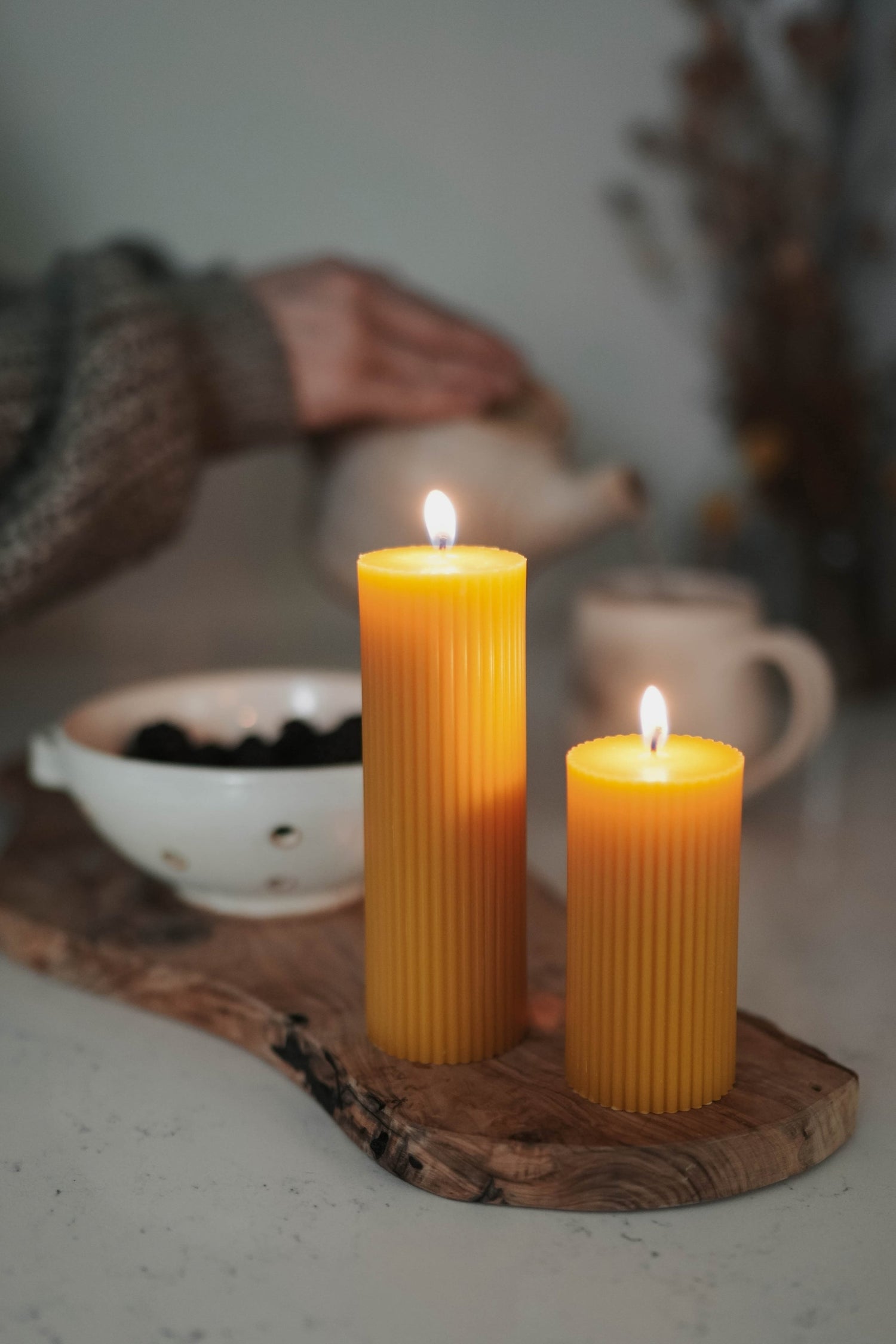Beeswax candles lit in the foreground while someone in a cozy knit sweater pours tea from a pot into a mug.